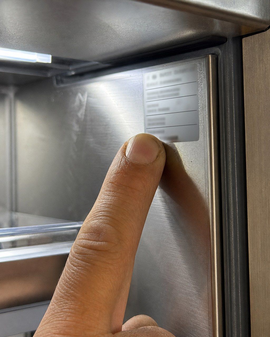 A technician’s thumb pointing at the silver model and serial-number sticker inside a Sub-Zero refrigerator cavity, with a slight glare on the stainless